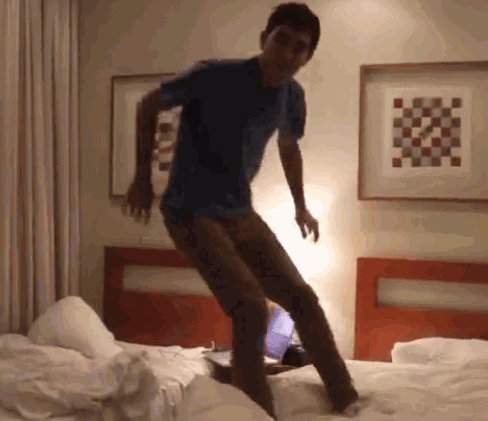 A guy jumps on top of a bed with his clothes on and gets underneath the bed covers with his clothes on top of the bed covers.