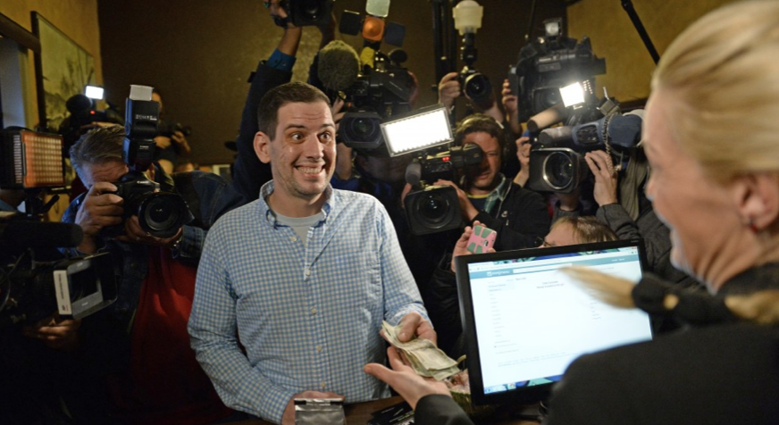 A man smiles while he purchases legal marijuana in Colorado. He hands over cash to a woman behind a desk as the media crew behind him take pictures.