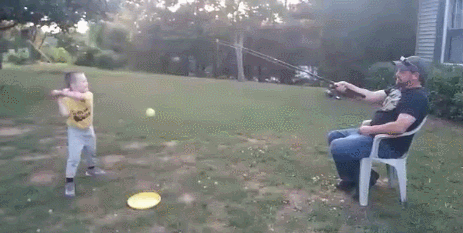 A lazy dad sits on a chair and uses a fishing rod to play baseball with his son.